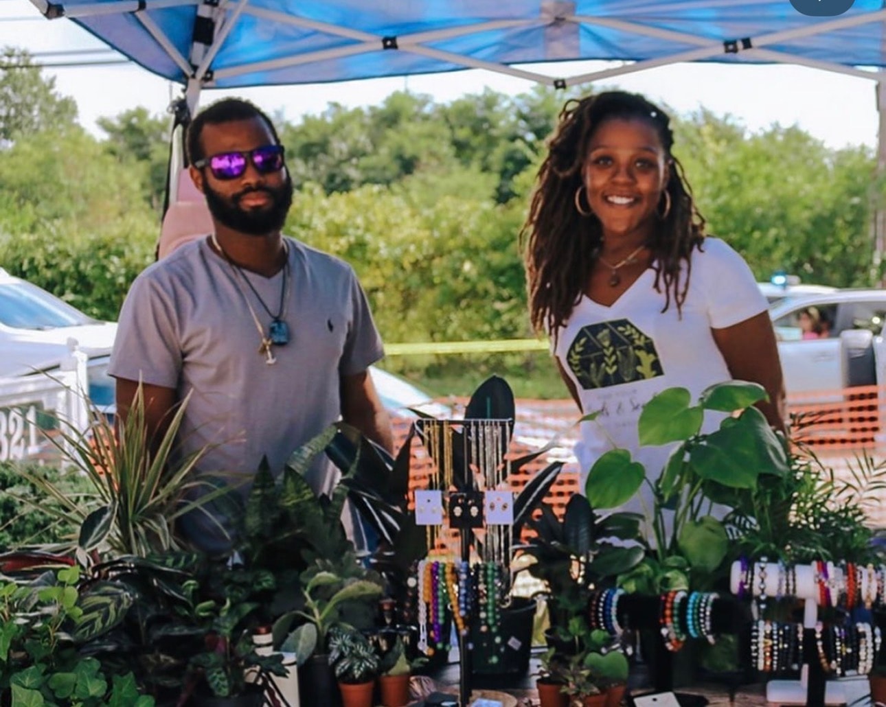 Two vendors smiling behind a market table full of plants and beaded jewelry under a blue tent