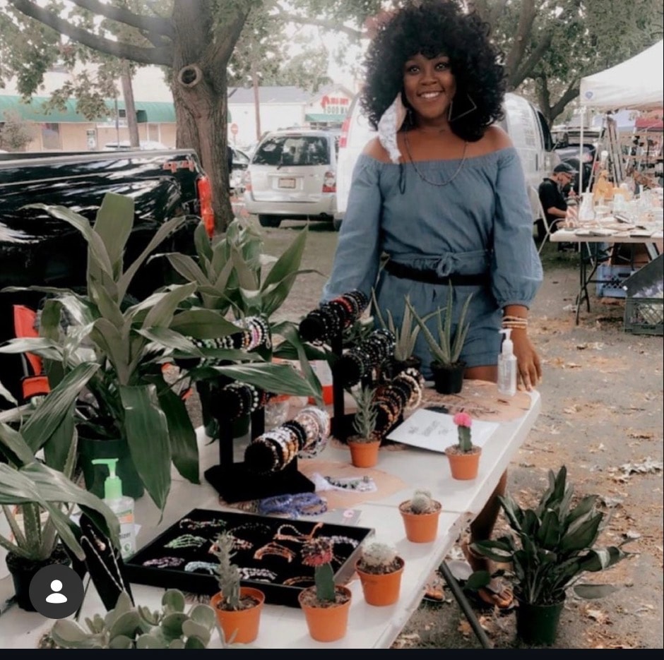 Vendor smiling at outdoor market with bracelets and small potted plants on display