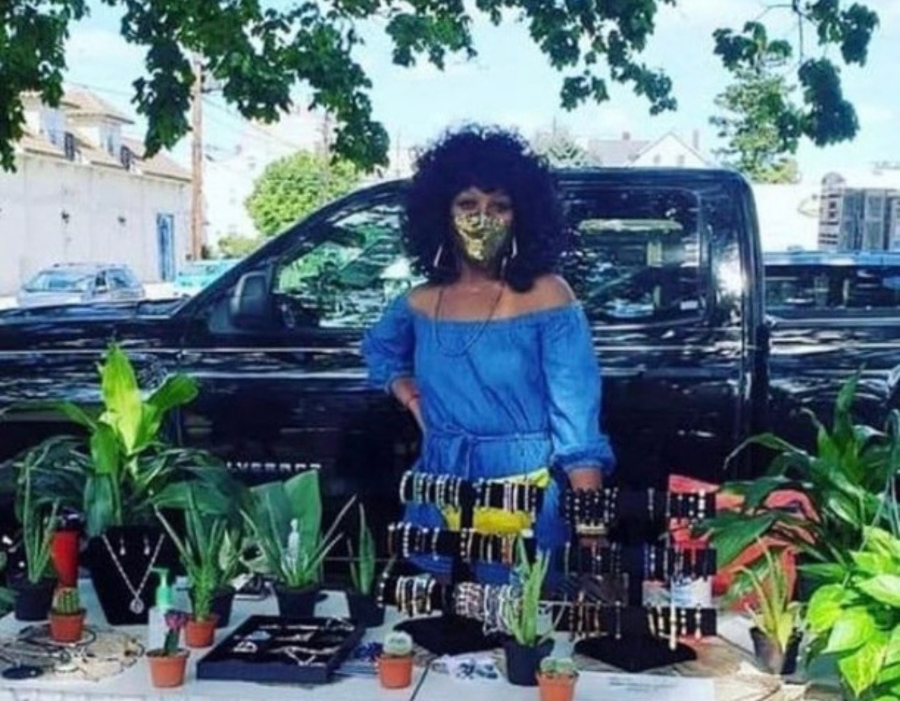 Vendor standing behind outdoor market table with plants and beaded jewelry displays