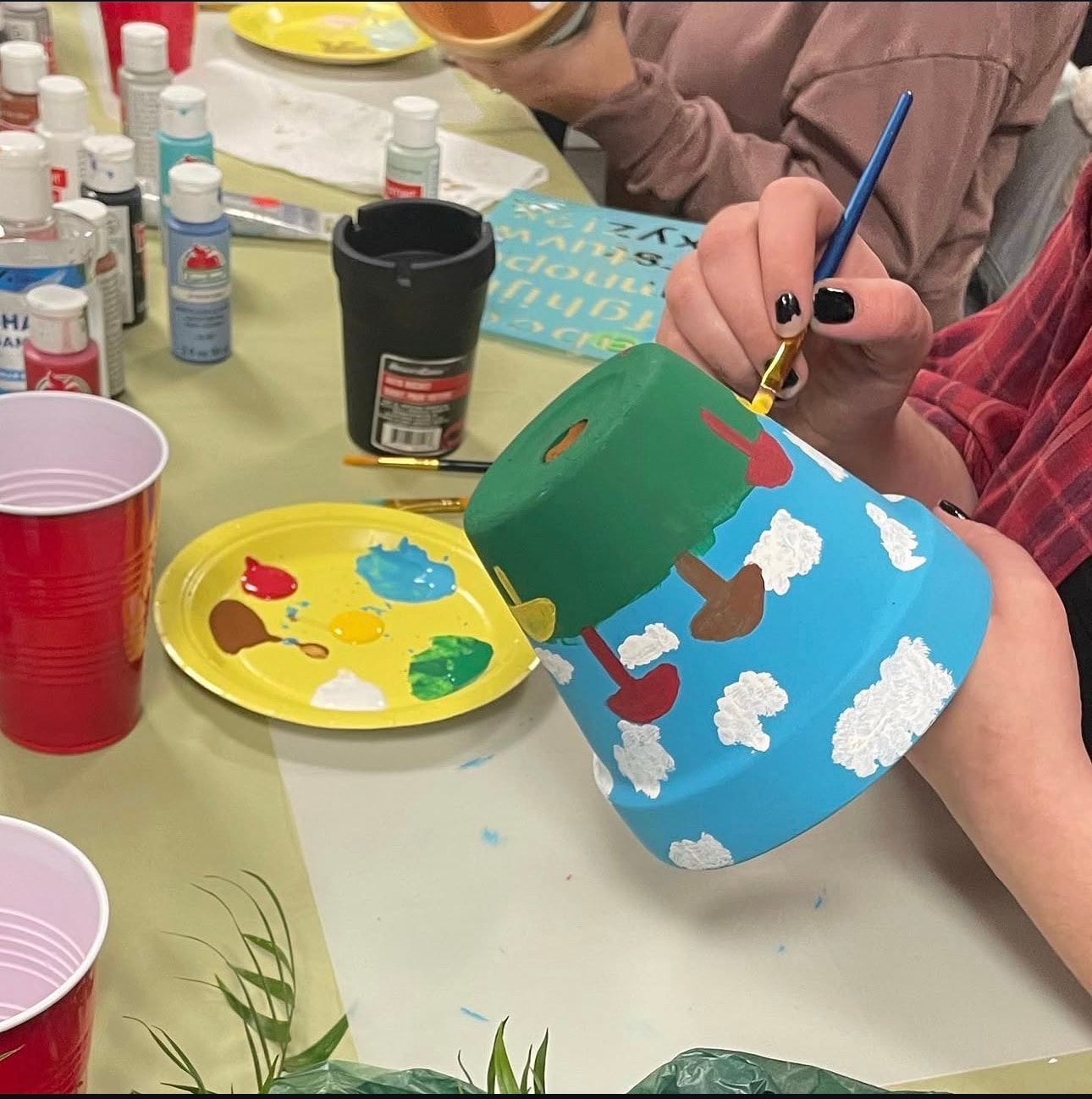 Close-up of hands painting a sky-and-clouds design on a terracotta pot at a workshop table