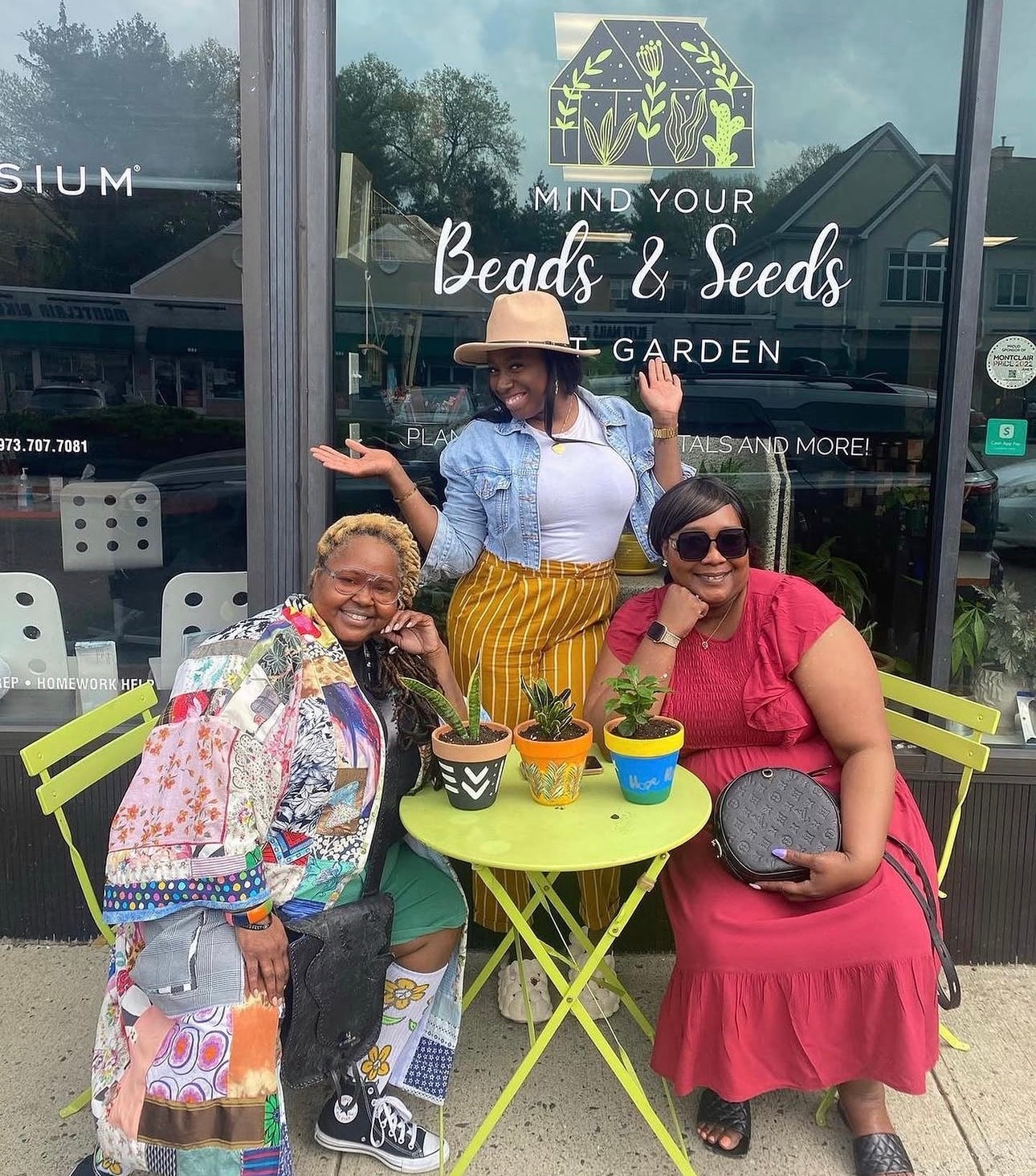 Three women posing with painted pots in front of the Mind Your Beads & Seeds storefront window